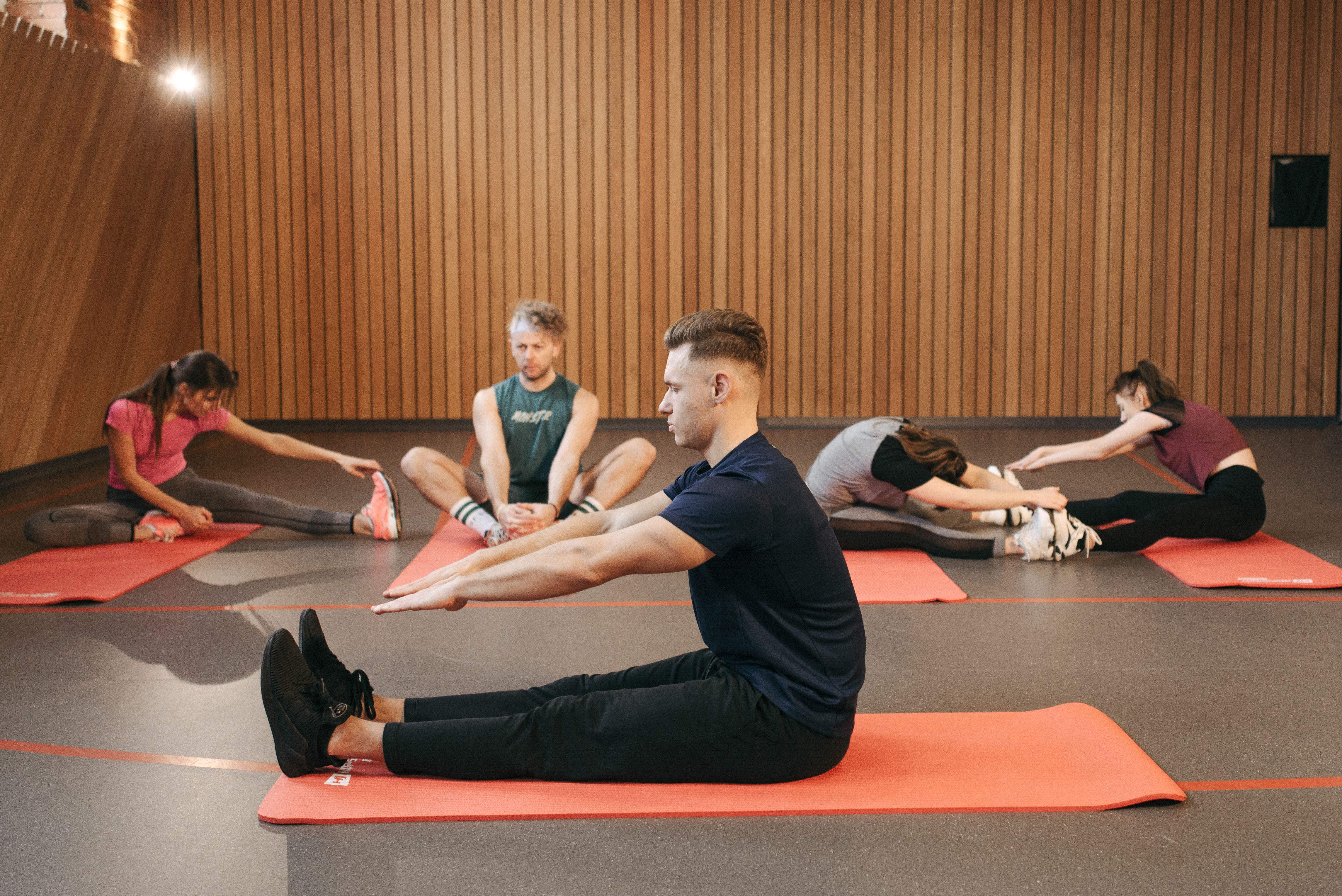 Five adults stretch while seated on orange exercise mats in a wood-paneled studio.
