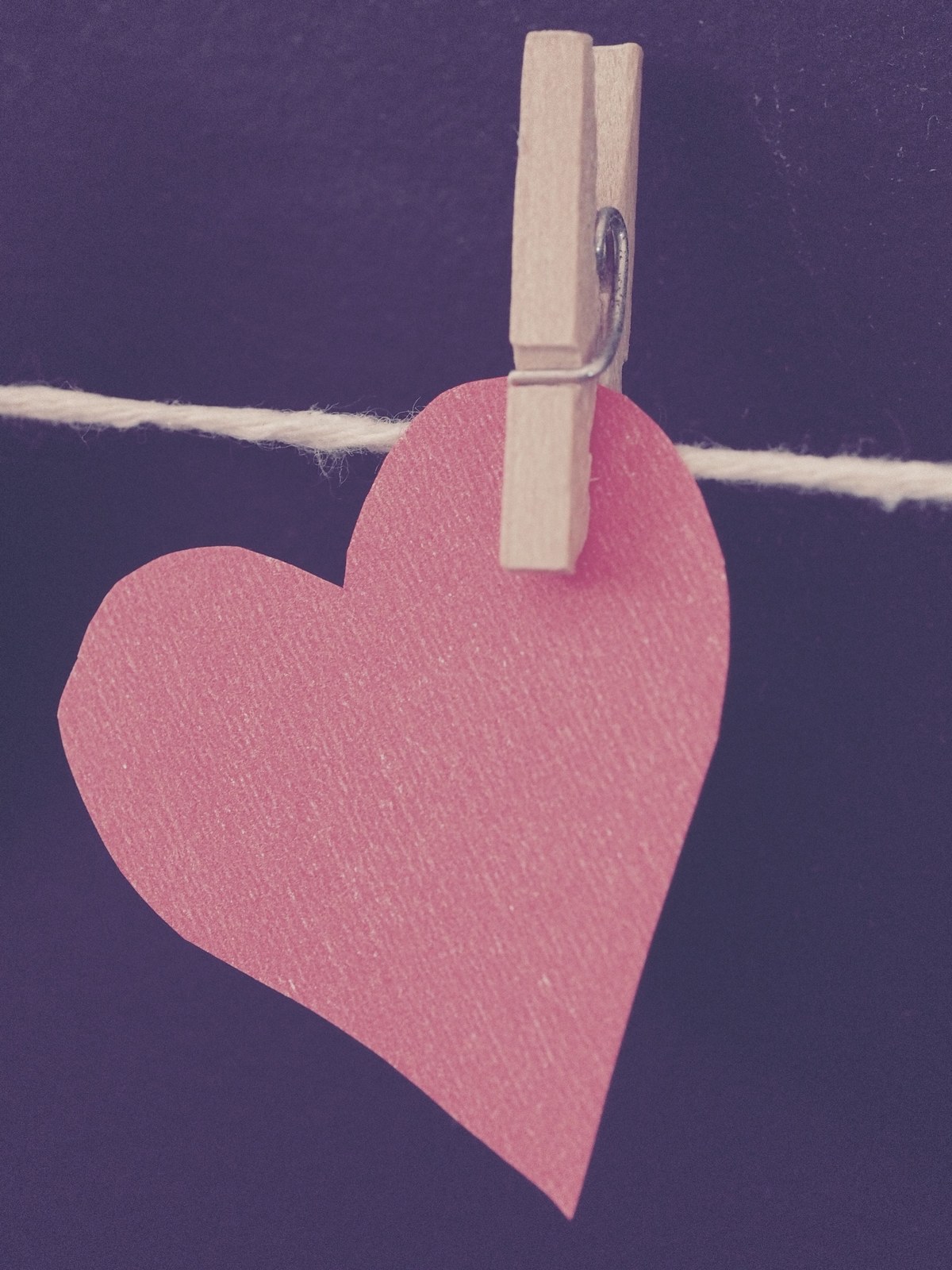 A pink-red paper heart hangs from string with a wooden clothespin against a dark blue-gray background.