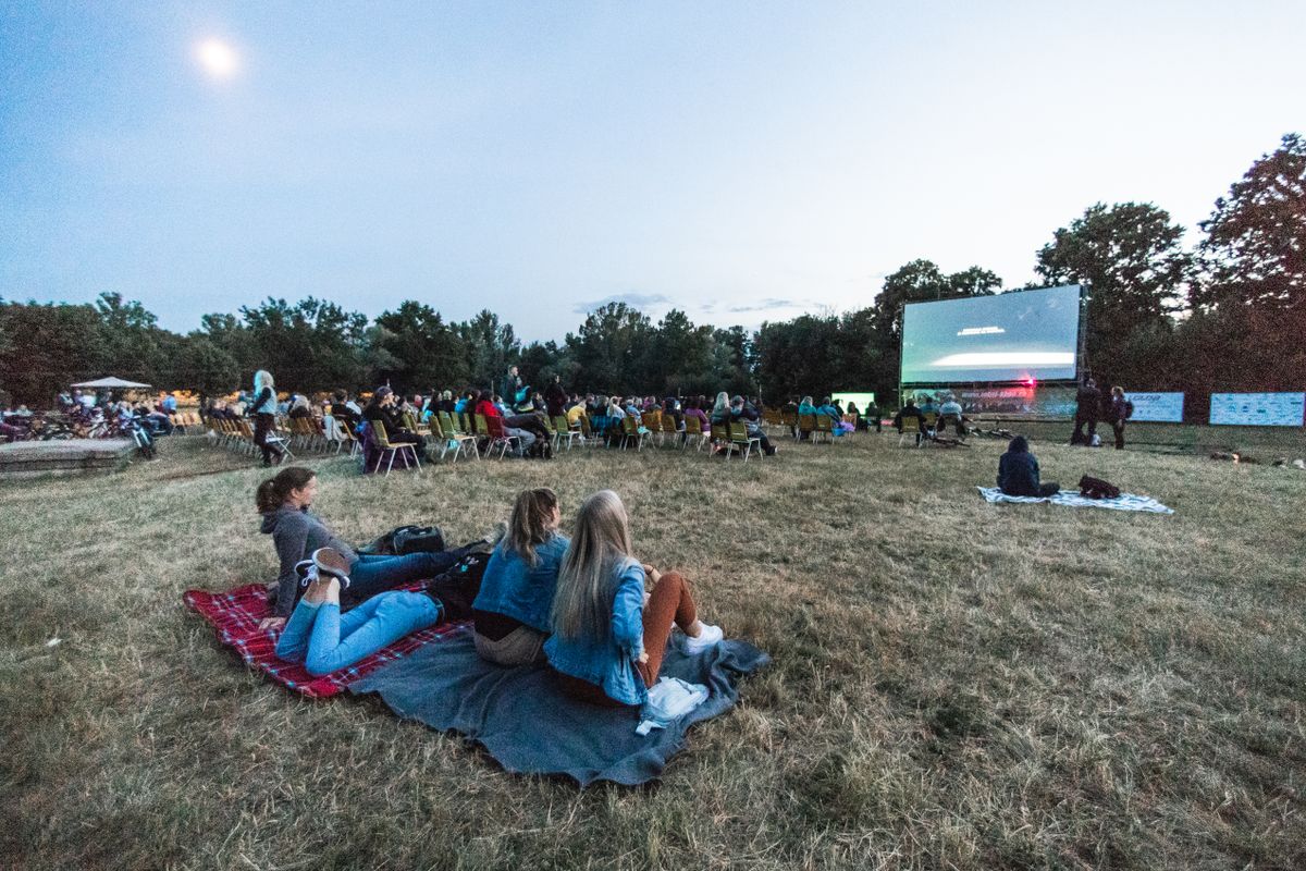 People sit on blankets and chairs facing an outdoor screen on a lawn at dusk.