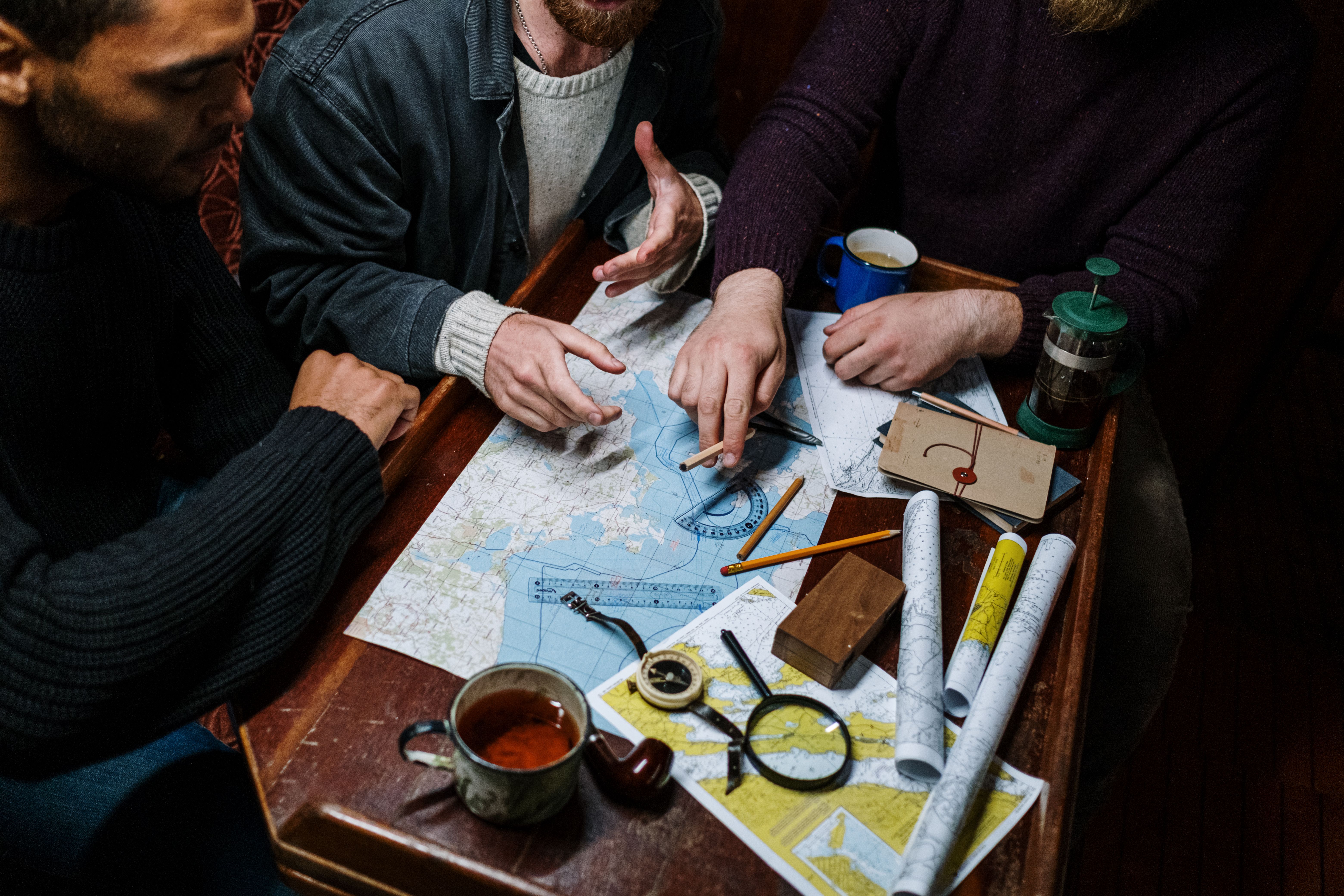 Three people study paper navigation charts around a wooden table with pencils, a compass and a magnifying glass.