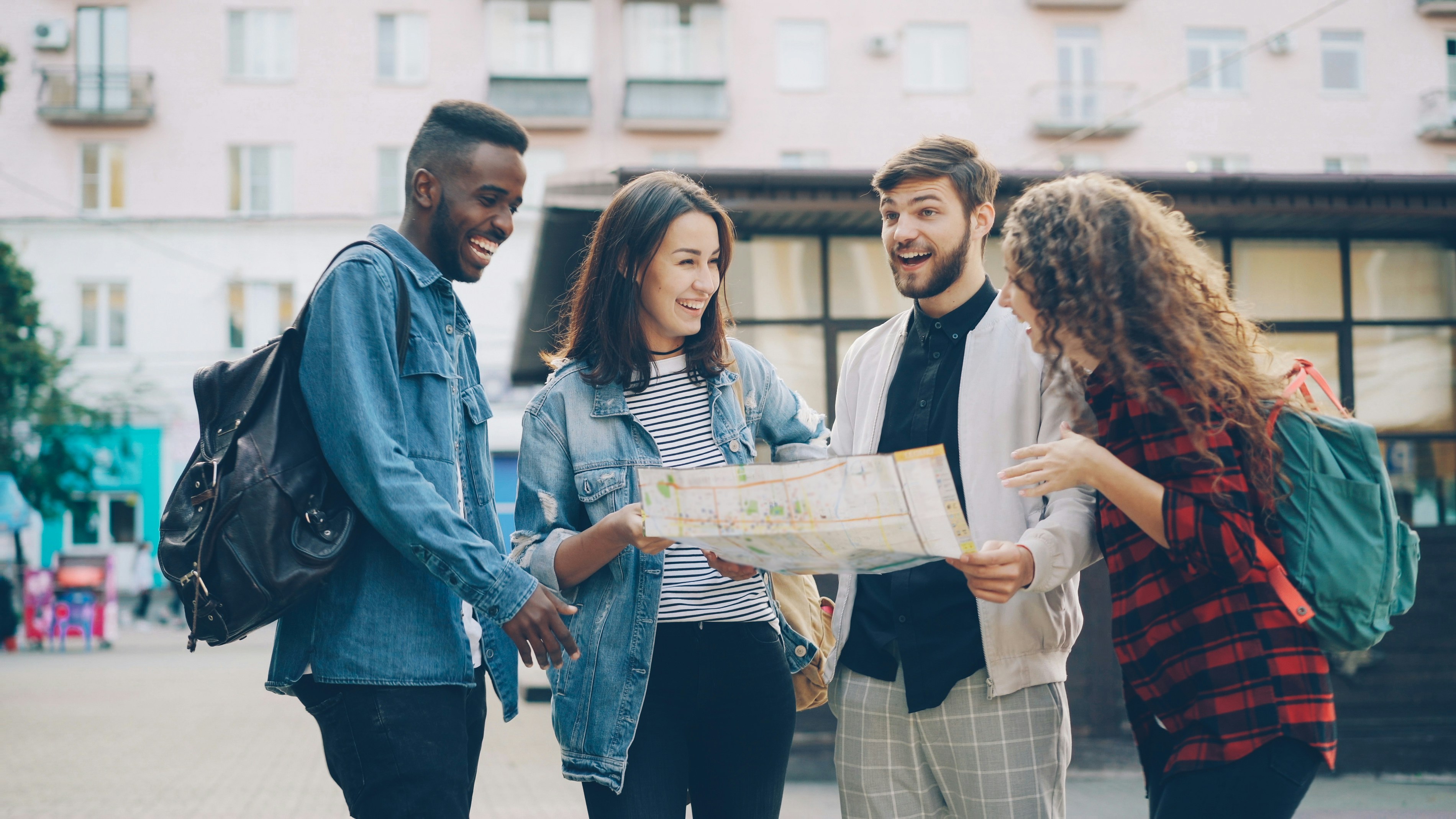 Four people with backpacks stand on a city street while two hold an unfolded paper map.