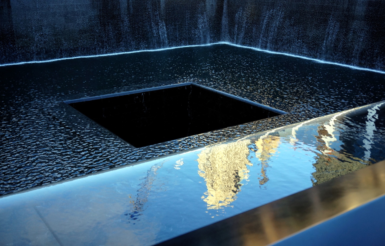 Water flows down the walls of a reflecting pool toward a square central opening at the National September 11 Memorial.