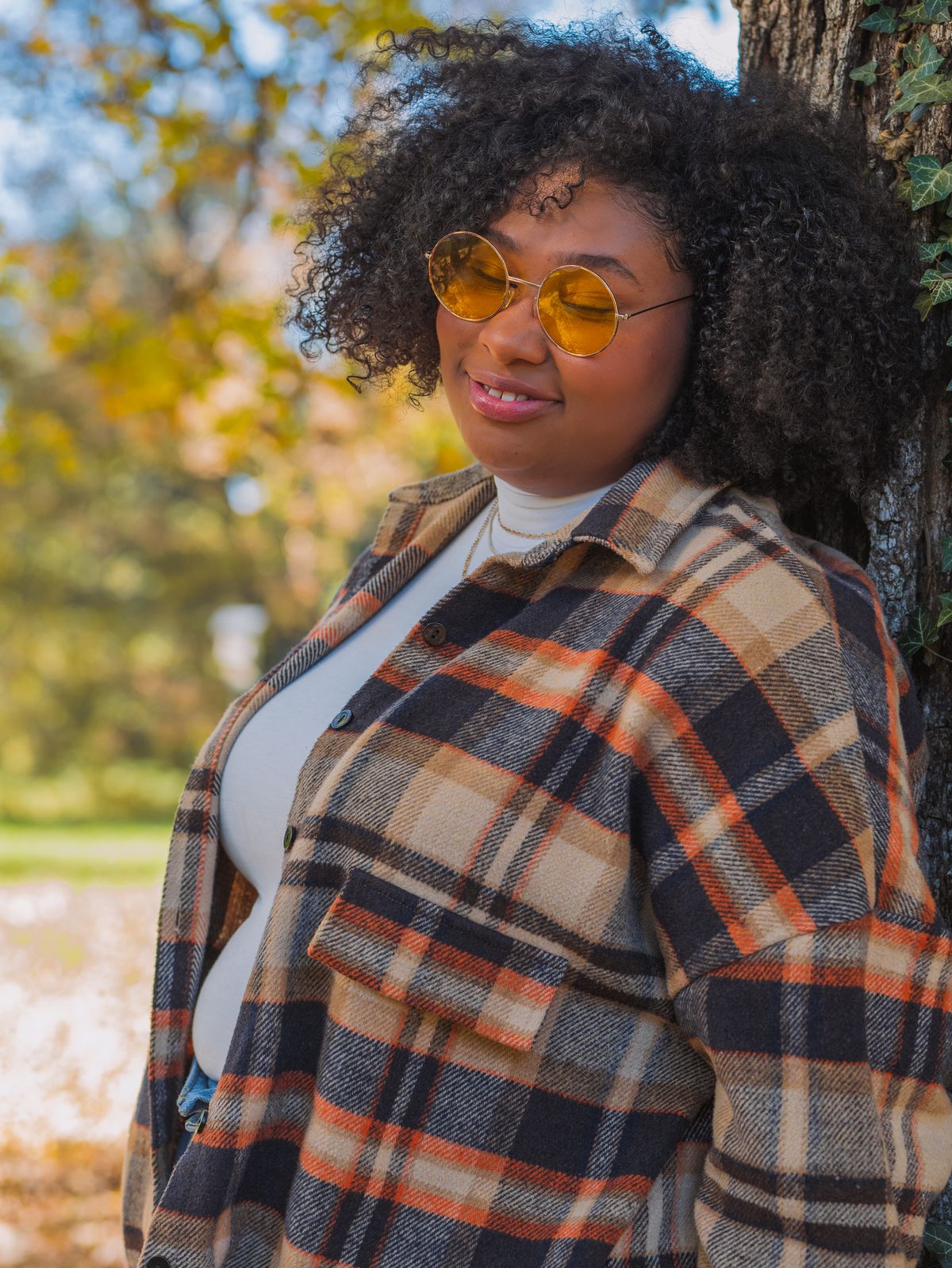 A woman in a plaid overshirt and yellow-tinted sunglasses stands beside a tree.