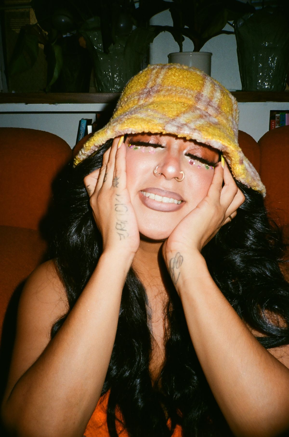 A woman wearing a yellow plaid bucket hat rests her hands against her cheeks.