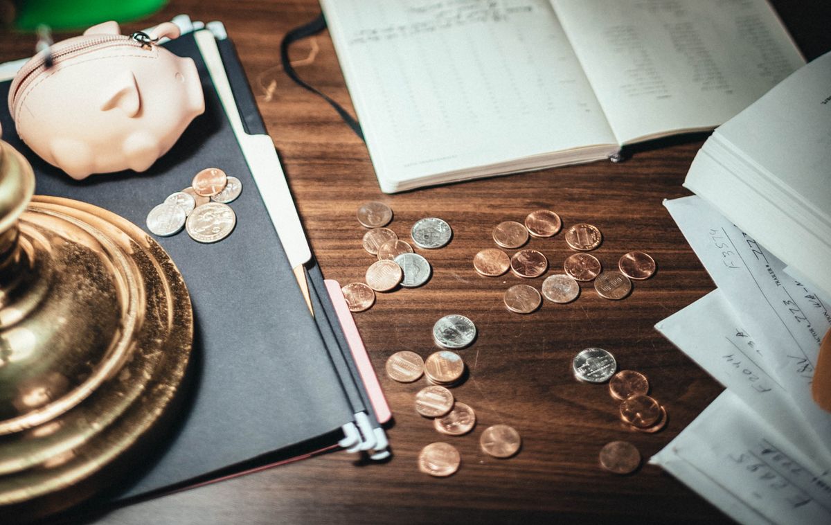 Coins lie on a wooden desk beside notebooks, papers and a pig-shaped coin purse.