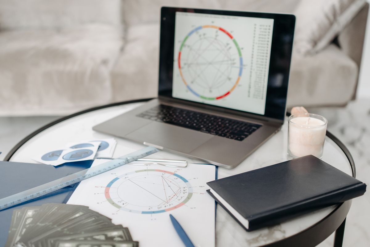 A laptop displays an astrology chart beside paper charts, cards and a notebook.