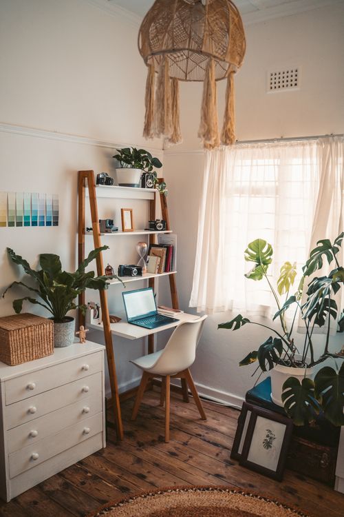 A small desk and chair sit beside a curtained window, surrounded by houseplants and shelves.