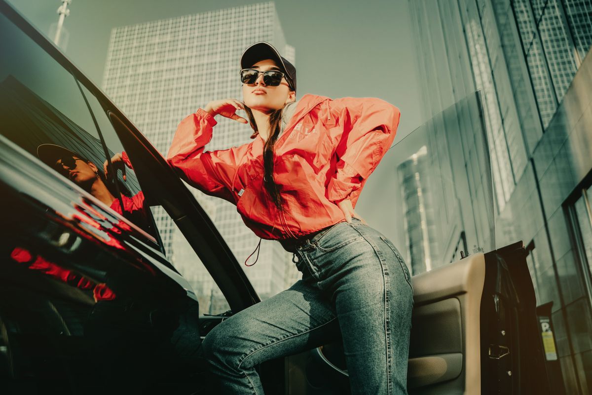A woman in a red windbreaker, blue jeans and black cap poses beside a car.