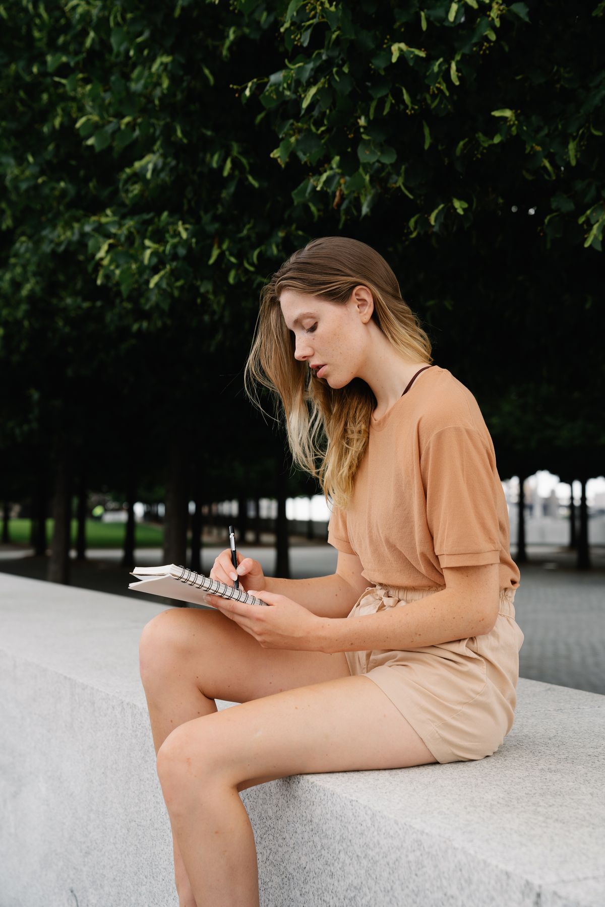 A woman sits on a low wall under trees and writes in a spiral notebook.