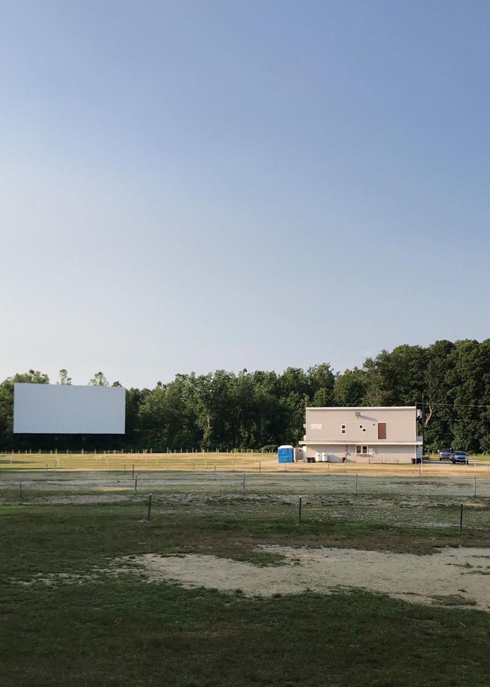 A blank drive-in screen and concession building stand beyond a grassy parking area.