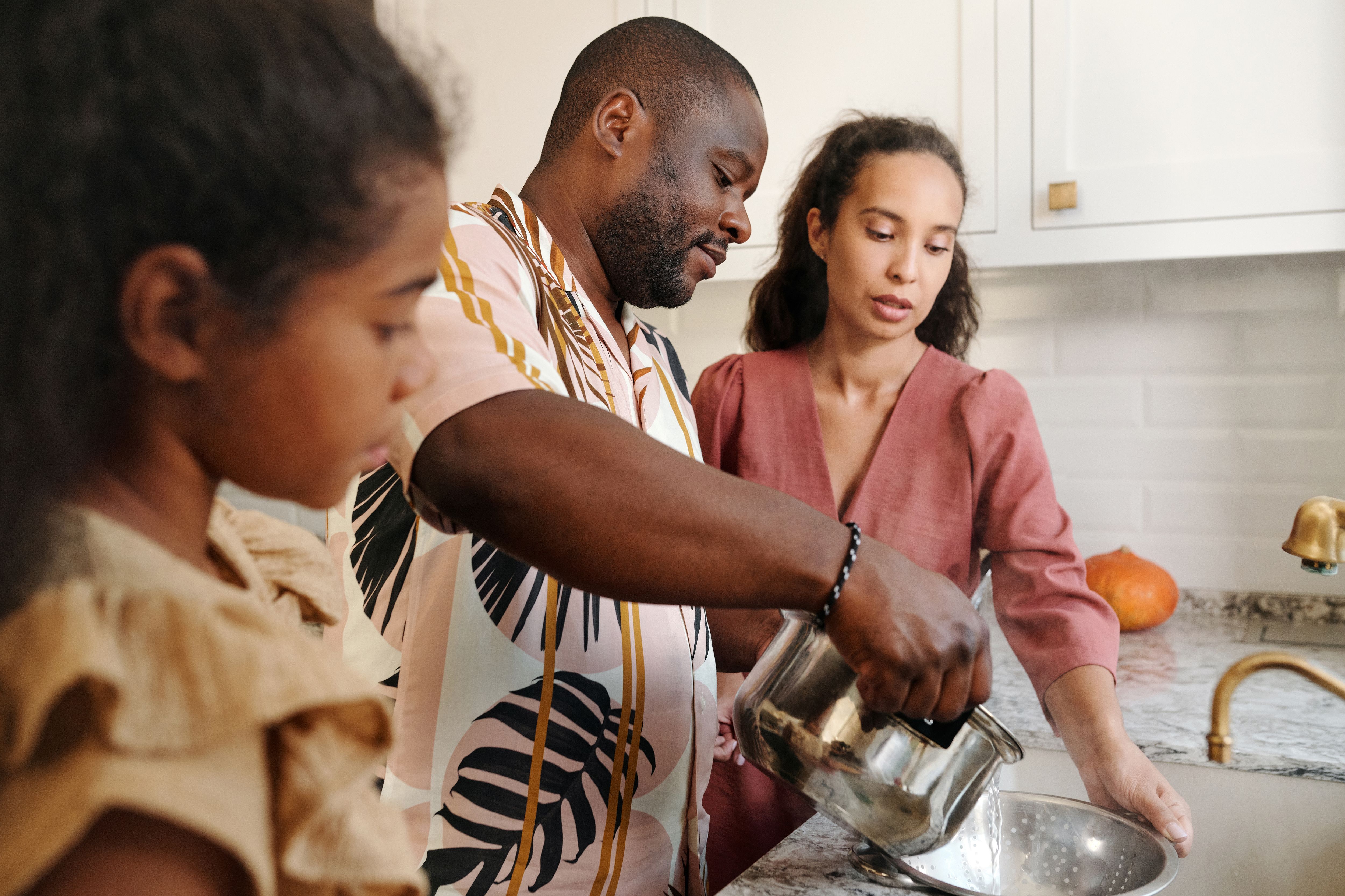 Two adults and a child prepare food at a kitchen sink, with a pot held above a colander.