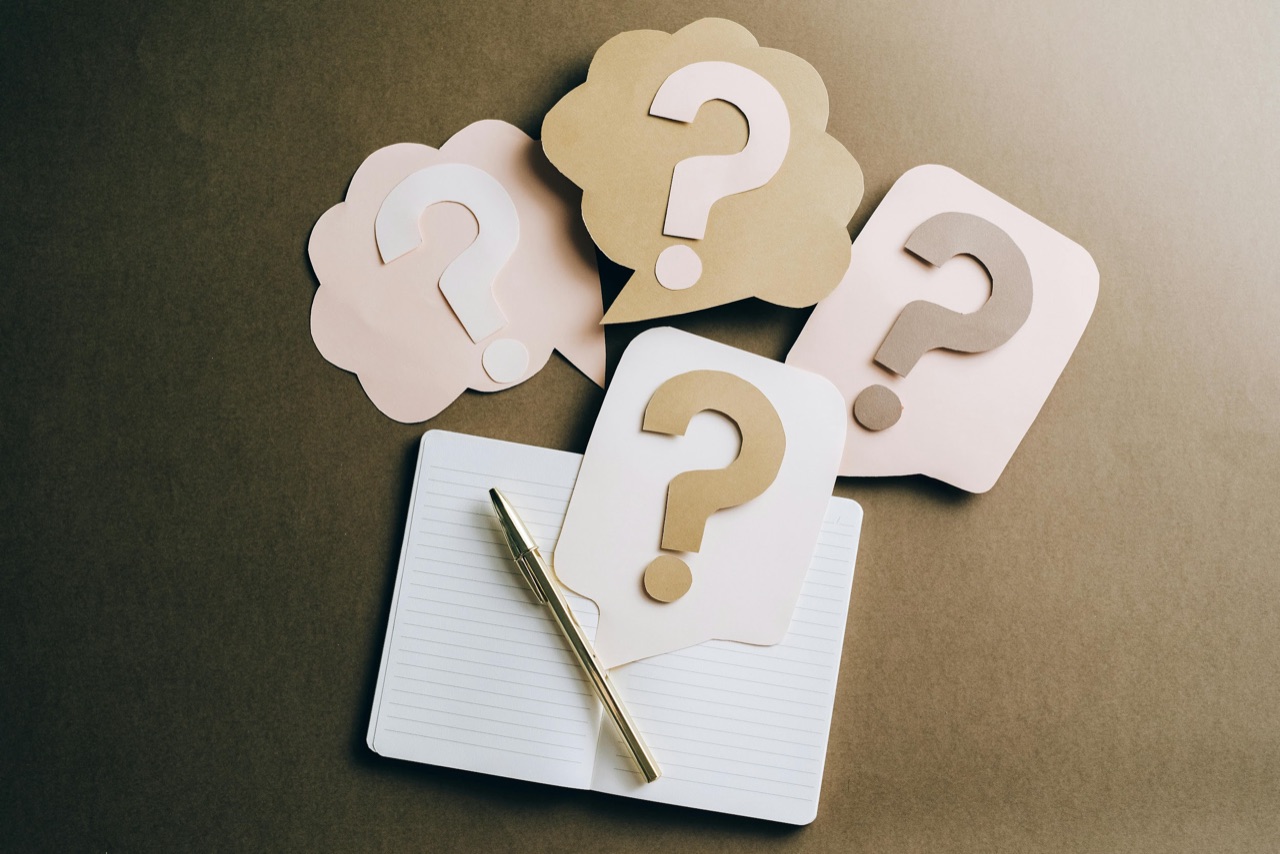 Four paper question-mark bubbles sit beside an open lined notebook and a gold-colored pen.