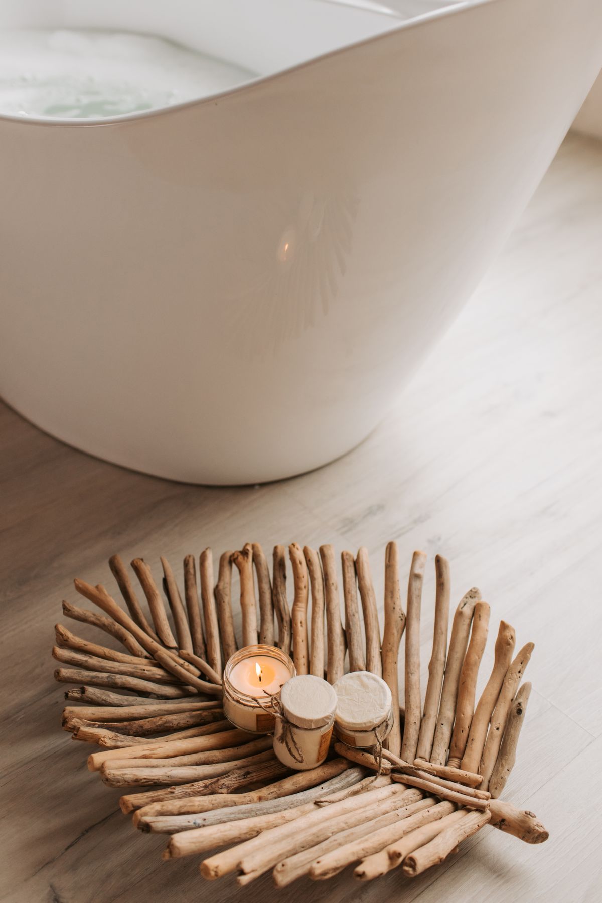 A white bathtub stands beside a wooden chair holding a lit candle and two covered containers.