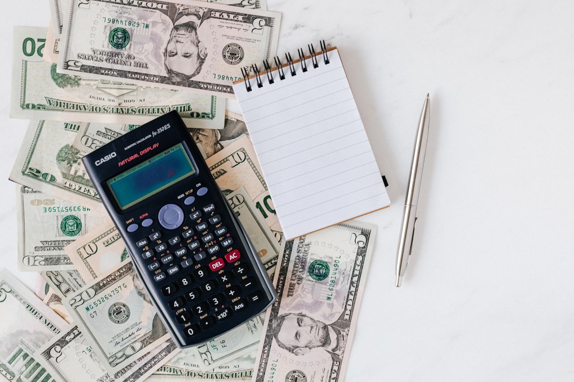 A calculator and blank notepad rest on US dollar bills beside a pen.