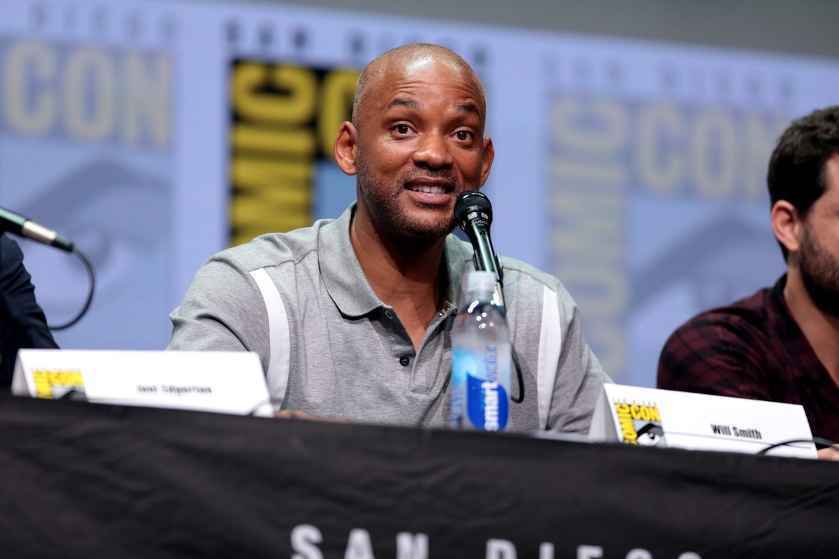 Will Smith sits behind microphones at a convention panel.