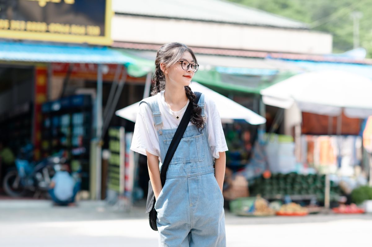 A woman wearing pale blue denim overalls over a white shirt stands outdoors.
